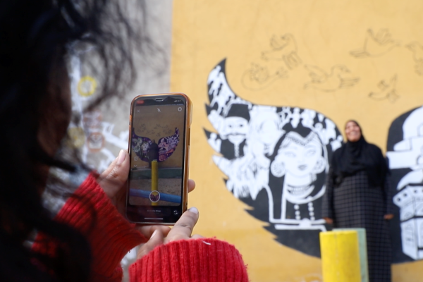 Image of a person looking through the camera app on a mobile phone at another person leaning against a wall with a mural in the background
